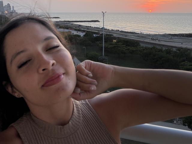 Gabrielafoxxy with her eyes closed, smiling softly, has a cute face and is wearing a sleeveless ribbed top, showing her bare shoulders and a slight tan. Sunset in the background.