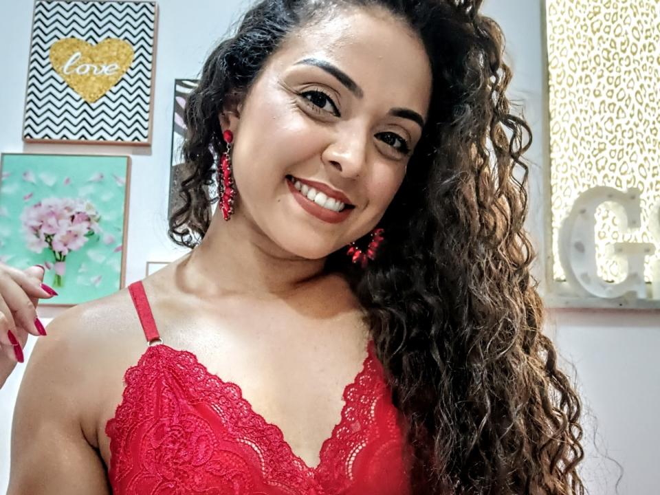 LaisMiguel, com cabelos castanhos cacheados, usando um sutiã de renda vermelho, sorrindo, com brincos vermelhos, unhas vermelhas, mostrando os seios e um rosto bonito. Ela é sexy e confiante..