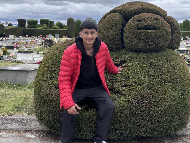 KaelNova, with short black hair, wearing a red puffy jacket and black clothes, sits on a giant, green, bushy monkey butt-shaped thingy in a cemetery.