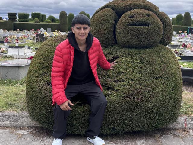 KaelNova sits on a massive green bush shaped like a fat face, wearing a red puffy jacket, black hoodie, and black pants, with white sneakers. Overcast sky.