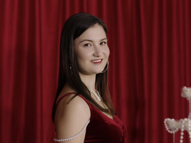 YungSifford tem cabelos longos e pretos, pele clara e um sorriso adorável. Ela está vestindo um vestido de veludo vermelho, um colar de pérolas e brincos, com uma pulseira de pérolas no braço direito..