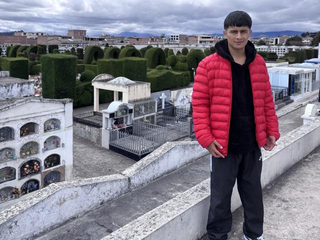 KaelNova, standing on a rooftop, wearing a red puffy jacket, black hoodie, and black pants, with short black hair, looking slightly to the side, in a cloudy outdoor setting.