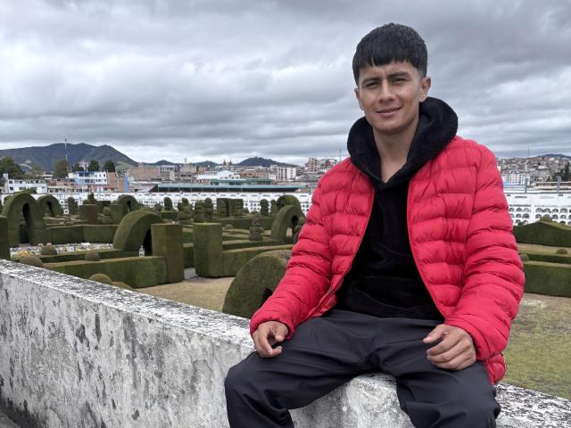 KaelNova, a young dude with short black hair, wearing a red puffy jacket, black hoodie, and black pants, sitting on a concrete wall, smiling at the camera.