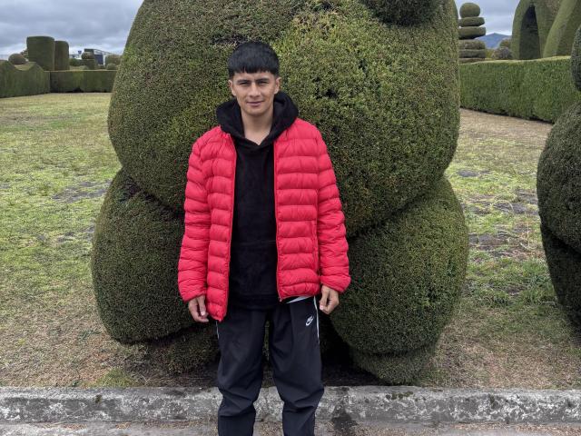 KaelNova, with short black hair, wearing a red puffy jacket and black hoodie, stands in front of a giant green topiary, in black pants, on a grassy area.