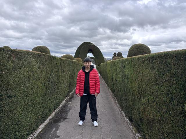 KaelNova stands in a narrow, green hedge-lined path, wearing a red puffer jacket, black hoodie, black pants, white sneakers, and a grey beanie, under a cloudy sky.
