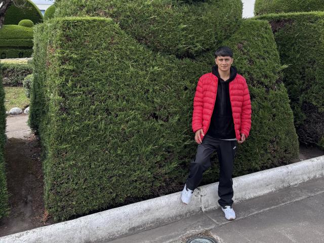 KaelNova stands against a green hedge, wearing a puffy red jacket, black shirt, black pants, and white sneakers. She's leaning on a concrete curb, smiling slightly.