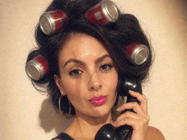 JakyKidman con cabello negro rizado, vestida con ropa roja y plateada, sosteniendo un teléfono negro, con lápiz labial rosa y aretes de aro dorados, mostrando una expresión de labios fruncidos..