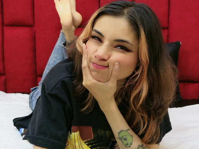 Amuneet with brown and blonde hair, black t-shirt, and tattooed arm, smiling with hands on cheeks, legs up, bare feet, red headboard, and white bed.