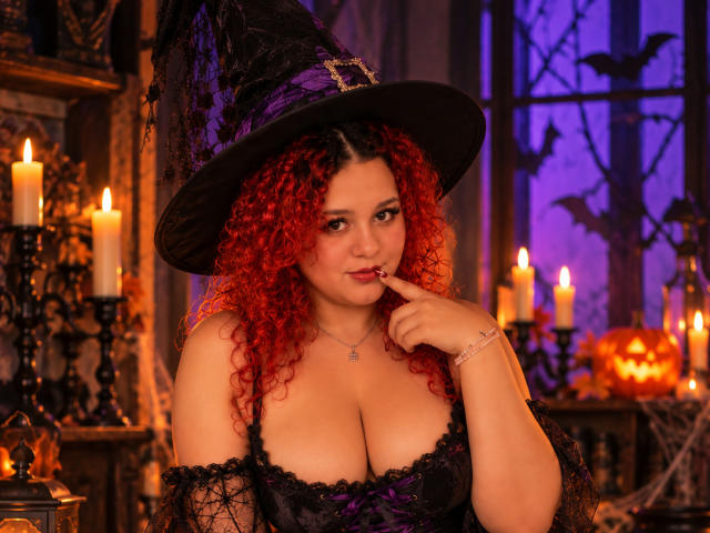 RosalieVittoria, avec des cheveux roux bouclés, un chapeau de sorcière noir, une robe en dentelle violette, un doigt sur les lèvres, montrant de grandes seins, entourée de bougies, d'une citrouille et d'un éclairage violet..