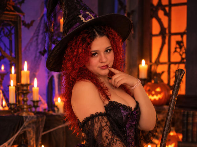 RosalieVittoria, con cabello rizado rojo, usando un sombrero de bruja negro, un corpiño de encaje morado y negro, y con un dedo en la boca, rodeada de velas y una calabaza tallada. Pechos, linda..
