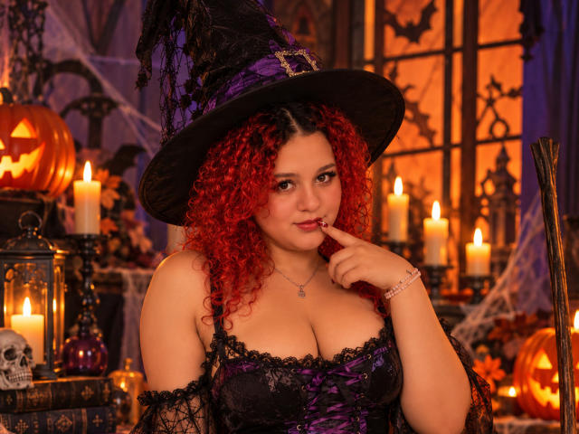 RosalieVittoria con i capelli rossi e ricci, indossa un corpetto di pizzo nero e un cappello da strega viola, con un dito in bocca, candele e zucche sullo sfondo, sexy, seno prosperoso, atmosfera di Halloween..