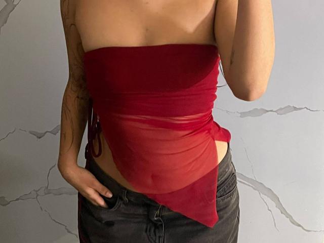 HannaThorne wearing a red, semi-transparent tube top that reveals her small tits and a tattoo on her left arm, with dark jeans and her right hand on her hip.