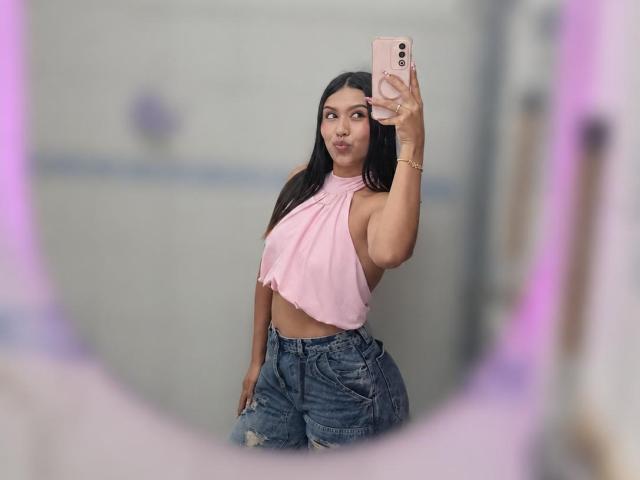 ZammySanz macht ein Selfie, trägt ein pinkes Halstopp und zeigt ihr großes Hinterteil in hoch taillierten, zerrissenen blauen Jeans. Sie hält ein pinkes Handy und hat langes, schwarzes Haar..
