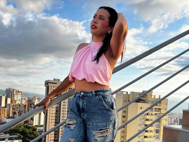 ZammySanz, con capelli neri, una canottiera rosa e jeans blu strappati, appoggiata a una ringhiera, con una mano tra i capelli, sullo sfondo si vede il panorama della città, il cielo è nuvoloso, illuminato dal sole, e guarda verso l'alto..
