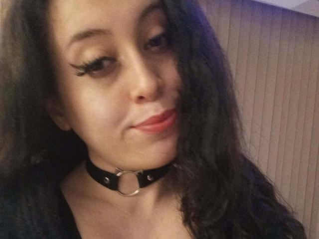KendraSimons a de longs cheveux noirs, porte un collier noir avec une bague, et a du rouge à lèvres avec des cils épais, souriant légèrement à la caméra..