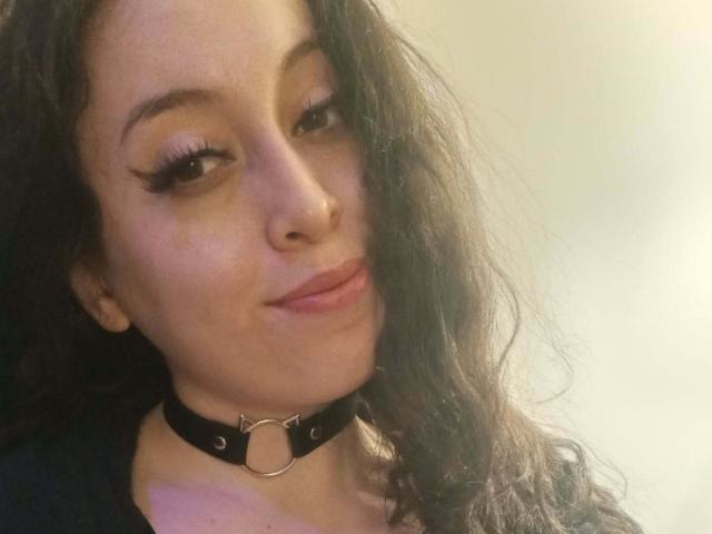 KendraSimons, avec ses cheveux noirs ondulés, porte un collier noir avec un anneau métallique, a de longs cils et un léger sourire, laissant apparaître son nez et ses lèvres roses..