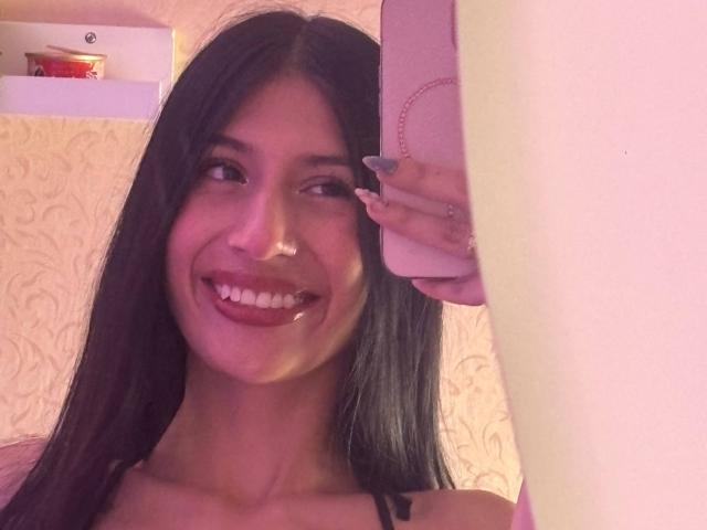 SophieBeaumont with long black hair, smiling with tongue out, holding pink phone, showing off nails, in a pink-lit room. Boobs not visible, focus on face.