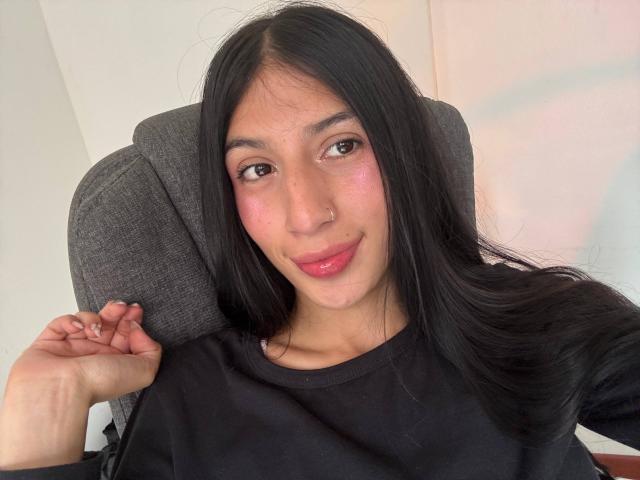 SophieBeaumont has long black hair, pink blush on her cheeks, and a slight smile. She's wearing a black shirt, sitting in a gray chair, with her right hand raised.