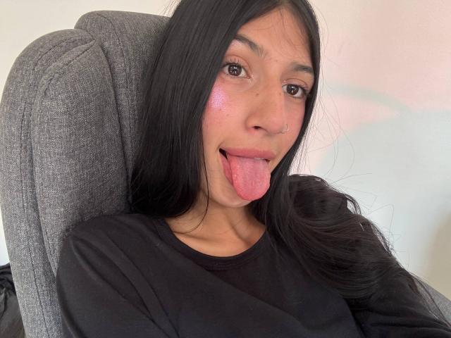 SophieBeaumont with long black hair, brown eyes, pink flushed cheeks, black shirt, nose ring, tongue out, sitting in gray chair. Boobs not visible, ass not visible, pussy not visible.