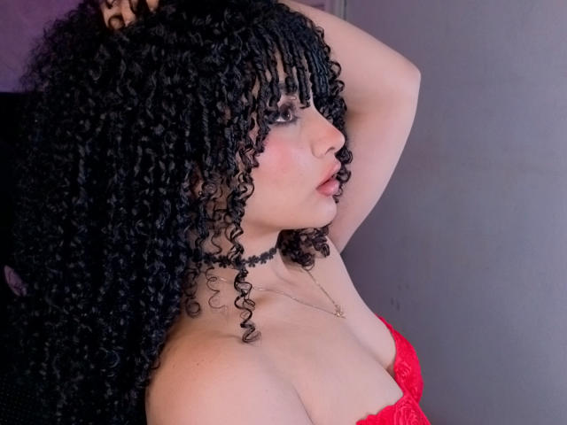 VanessaBlake with curly black hair, red lace top, pale skin, black choker, arm raised, side profile, showing her tits, looking to the right, subtle makeup, confident expression.