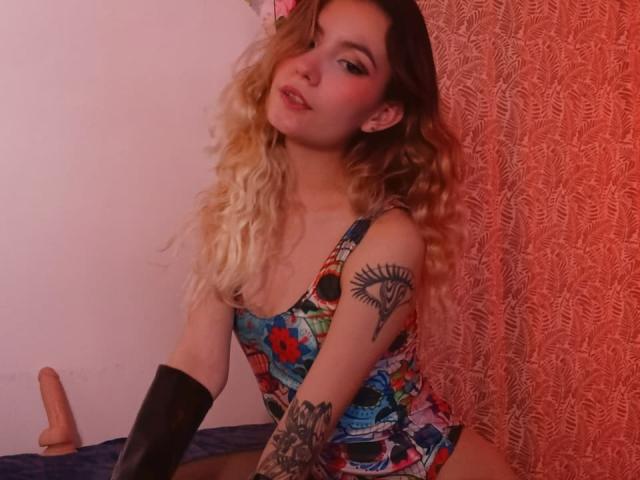 AlanisBlanc with wavy blonde hair, wearing a colorful, patterned, one-piece, showing a tattoo on the right arm, black elbow gloves, and a pinkish-red curtain background.
