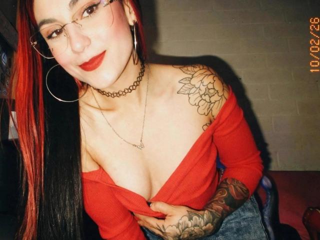 Amy, mit roten und schwarzen Haaren, Brille, rotem Top mit einem Schulter frei, Choker und Tattoos, zieht ihr Top herunter, um ihre Brüste zu zeigen. Datumsstempel in der Ecke..