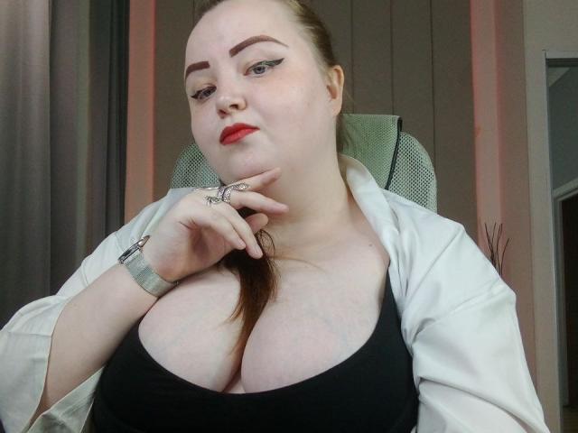 MissEmmar, com pele clara, batom vermelho e uma camiseta preta, mostrando seus seios grandes, cabelo castanho em um rabo de cavalo, roupão branco, anel e pulseira no pulso, olhando para a câmera..