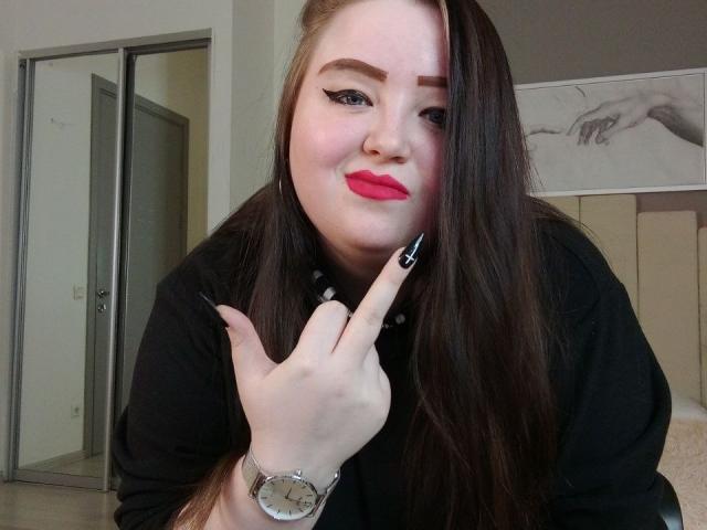 MissEmmar with long brown hair, black eyeliner, red lipstick, and black nails, pointing at her face with a finger, wearing a black shirt and a silver watch.