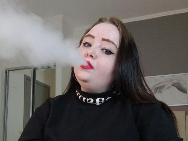 MissEmmar, met bleke huid, donkerbruin haar en rode lippenstift, blaast een grote witte rookring. Ze draagt een zwarte top met een zilveren ketting als halsband..