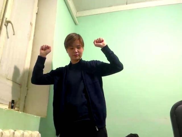 MarybelleDoerr stands with arms raised, wearing a black shirt and jacket, in a green-walled room. She has brown hair and a serious expression. Her ass is not visible.