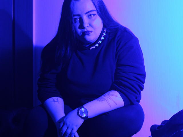 MissEmmar met blauw-paarse verlichting, in een hurkhouding, zwarte trui, lang bruin haar, polstattoo, horloge, zwarte broek, zelfverzekerde uitdrukking, donkere achtergrond, zichtbare tatoeage op de linkeronderarm..