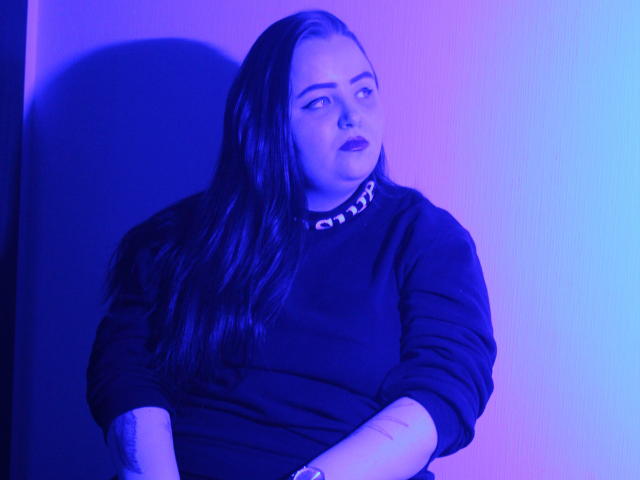 MissEmmar, com longos cabelos escuros, vestindo um suéter preto com uma gola de corrente, olhando para o lado, com uma luz neon azul e rosa projetando uma sombra sobre ela..