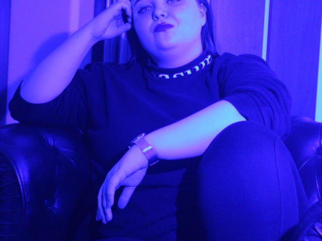 MissEmmar, avec une peau teintée de bleu, un haut noir et un pantalon noir, assise sur un canapé en cuir, tenant un téléphone à l'oreille, portant une montre, avec des lèvres boudeuses, un rouge à lèvres foncé et un anneau au nez..
