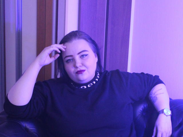 MissEmmar mit blauen Augen, dunklem Lippenstift und schwarzem Hemd, sitzt auf einem schwarzen Ledersofa, eine Hand am Kopf, hat ein Tattoo am rechten Unterarm, es ist violettes Licht, sie trägt eine silberne Uhr..