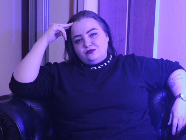 MissEmmar, con una mano sulla fronte, indossa un maglione nero con colletto decorato, seduta su un divano in pelle nera, illuminazione blu-viola, tatuaggio visibile sul braccio sinistro..