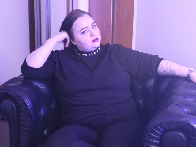 MissEmmar, con la pelle chiara, i capelli scuri e il rossetto, indossa una maglia nera a collo alto e pantaloni, è seduta su un divano in pelle nera, con una mano sulla testa, mostrando un tatuaggio sul braccio..
