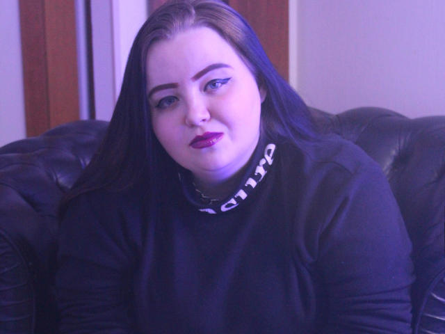 MissEmmar, com pele clara, cabelo azul escuro, batom roxo, gola alta preta e colar de corrente, sentada em um sofá de couro preto, olhando diretamente para a câmera, com um leve sorriso..