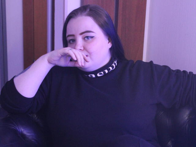MissEmmar, con piel pálida, cabello oscuro, ojos azules y un atuendo negro, mordiéndose el dedo, sentada en un sofá de cuero negro, usando un collar negro y blanco, con iluminación púrpura..