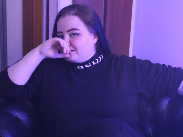 MissEmmar, com cabelos longos e escuros, um suéter preto e iluminação com tons de roxo, mordendo o dedo com um leve sorriso, sentada em um sofá de couro preto..