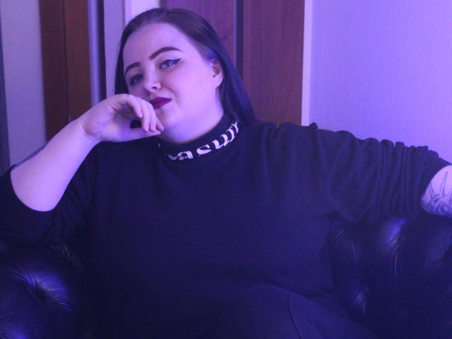 MissEmmar with dark hair, black turtleneck, and tattoo on right arm, sitting on black leather couch, resting chin on hand, blue-purple lighting, slightly smiling, looking at camera.