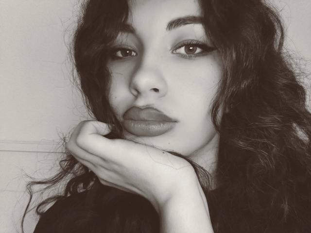 Foto em preto e branco de AliceKris com cabelo escuro e ondulado, apoiando o queixo na mão, olhando diretamente para a câmera com lábios cheios e levemente brilhantes e maquiagem discreta..
