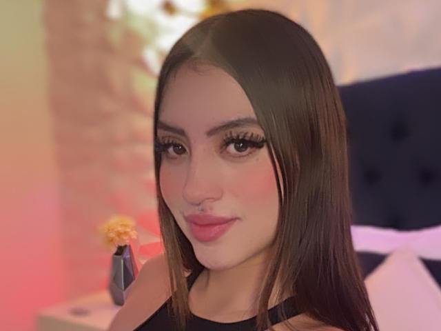 LunnaBlossom has long brown hair, full eyebrows, and wears heavy black eyeliner. She's looking at the camera with a slight smile, wearing a black top.