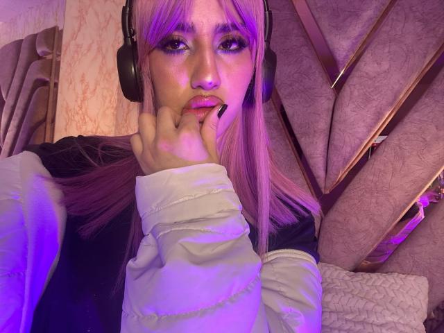 Elodie Roussee, con il viso illuminato di viola, cuffie nere, maniche bianche e gonfie, e capelli rosa, che si morde un dito, guarda la telecamera, indossando una maglietta nera..