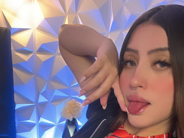 LunnaBlossom with dark hair, nose ring, and lip piercing, sticking out tongue, has long eyelashes, pink blush, and red shirt, with her hand near her cheek.