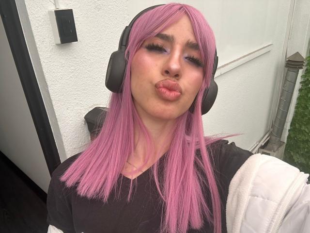 Elodie Roussee, con i capelli rosa, le cuffie nere, le labbra piene, una maglietta nera, una giacca bianca e un'espressione seria, è in piedi all'esterno. Il suo seno è piccolo e i suoi glutei non sono visibili..