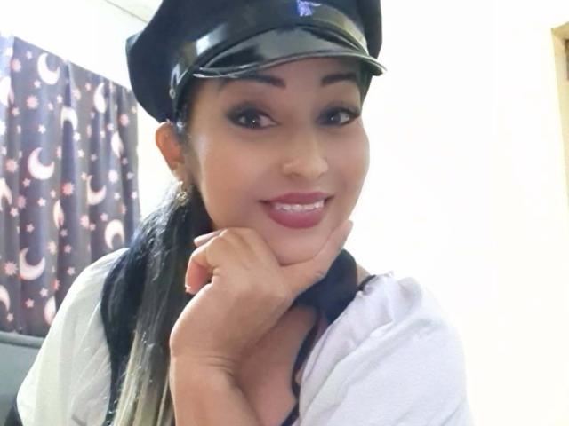 PielcanelaForU mit dunklem Haar, trägt eine schwarze Polizeimütze, ein weißes Hemd und eine schwarze Weste, lächelt mit rotem Lippenstift, die Hand ruht auf der Wange, sieht süß und sexy aus..