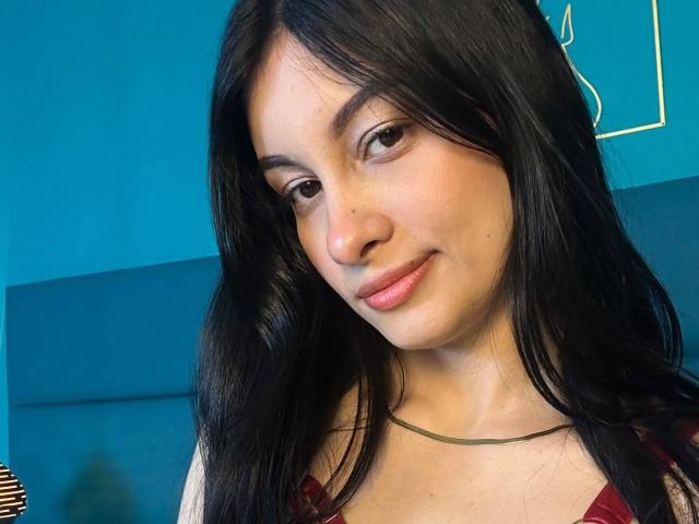DevineSimone has long black hair, brown eyes, and a slight smile. She's wearing a red top with a green necklace. Her tits are small and her skin is fair.