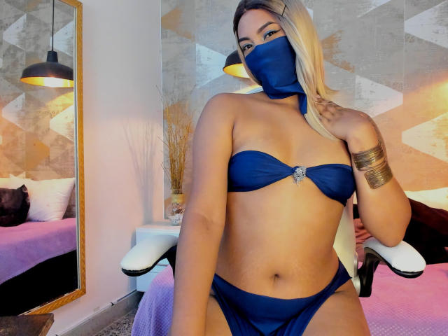 Lalila: Bruinharig meisje met blond haar, een blauw masker, een blauw bikinibroekje, een gouden armbandje, zittend op een stoel, die haar borsten en vulva laat zien, reflectie in een spiegel, warme verlichting in de slaapkamer, een roze bed, een beige muur..
