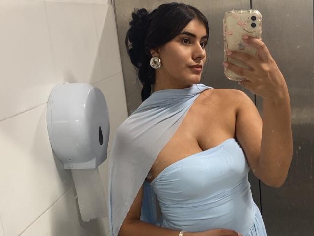 AlaraKey with dark hair in a bun, wearing a light blue, one-shoulder dress, showing her tits in a mirror selfie, holding a floral phone case, wearing round earrings.