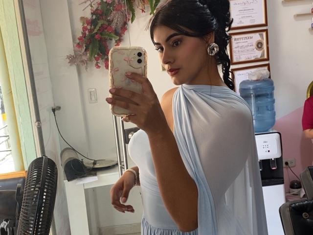 AlaraKey with dark hair in a bun, wearing a sheer blue top, showing her tits, holding a phone with a floral case, taking a mirror selfie in a room.
